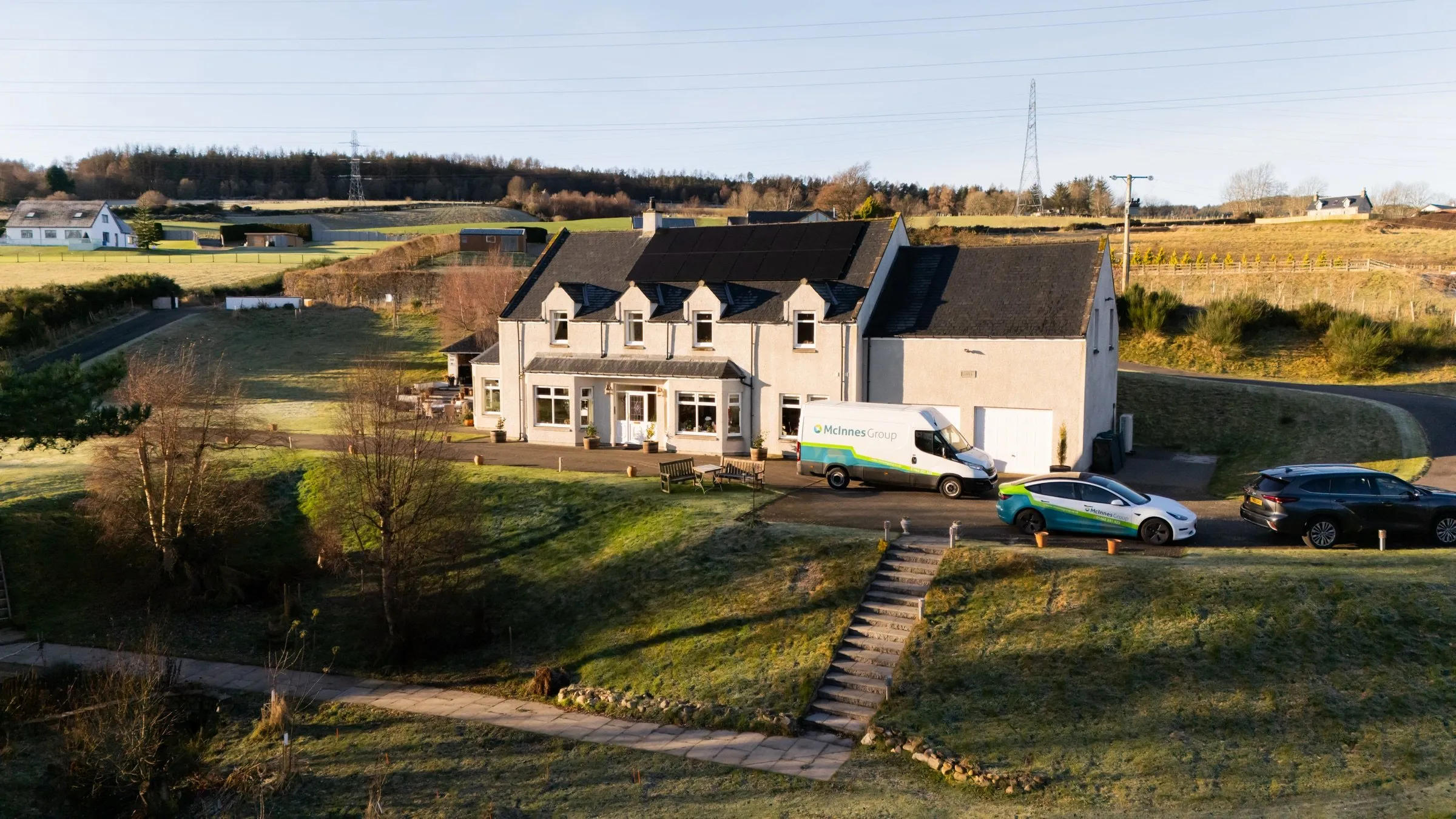 Drone view of a solar PV installation at a customer's home in Beauly, Inverness-shire, with McInnes Group vans and a Tesla Model 3 in the driveway.
